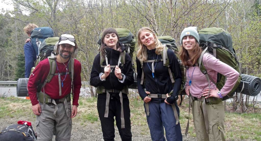 Four people wearing backpacks smile for a photo in a wooded area